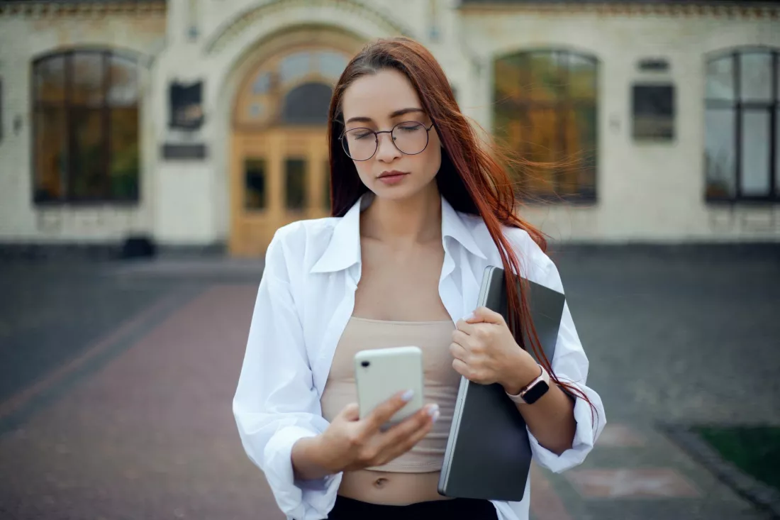 Studentka w okularach patrzy w smartfon przed budynkiem uczelni